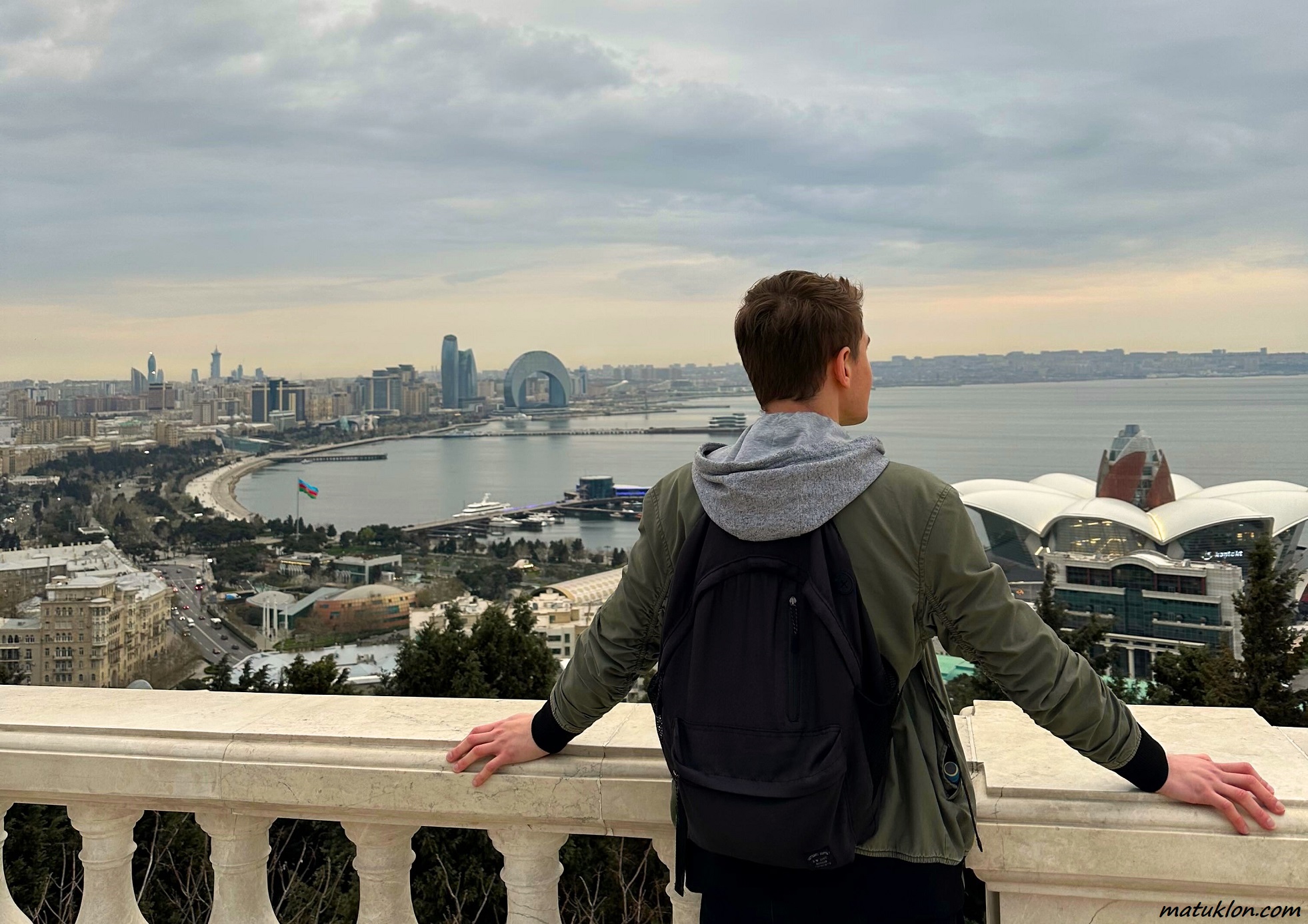 Man in green jacket and with a black backpack standing at a white marble balcony, looking at a bay and the city below