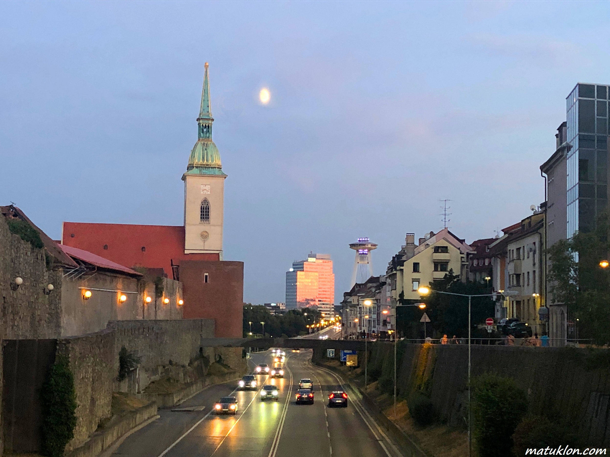 Church tower and modern buildings above a busy road with cars, set against grey sky at dusk with a moon