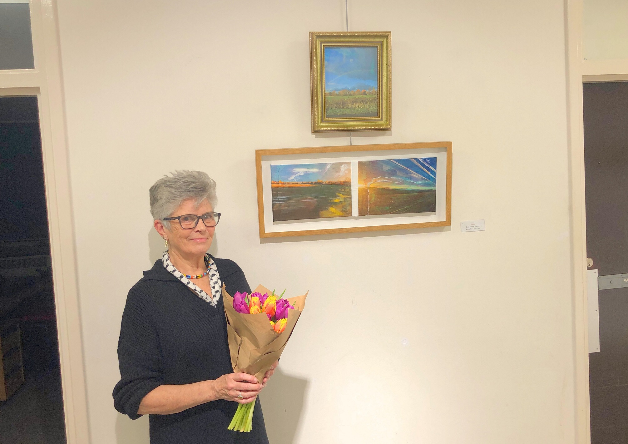 Woman dressed in black, holding a bouquet of flowers, standing next to artworks hung on a wall