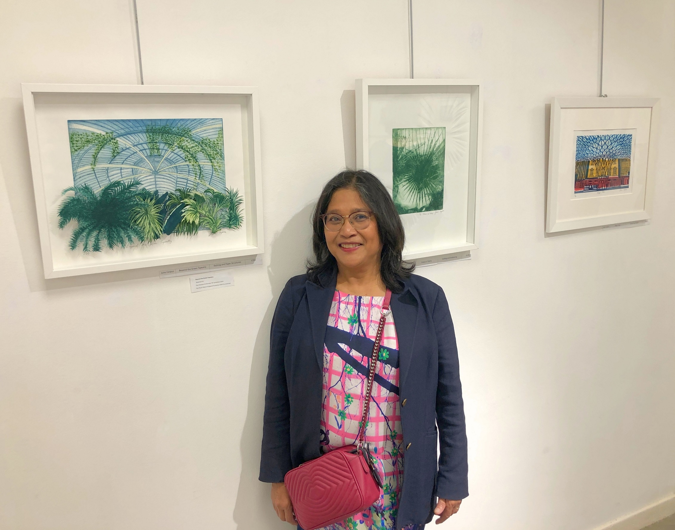 Woman wearing a colourful dress, a dark navy blazer and a dark pink handbag standing in front of a wall with two green-tinted artworks