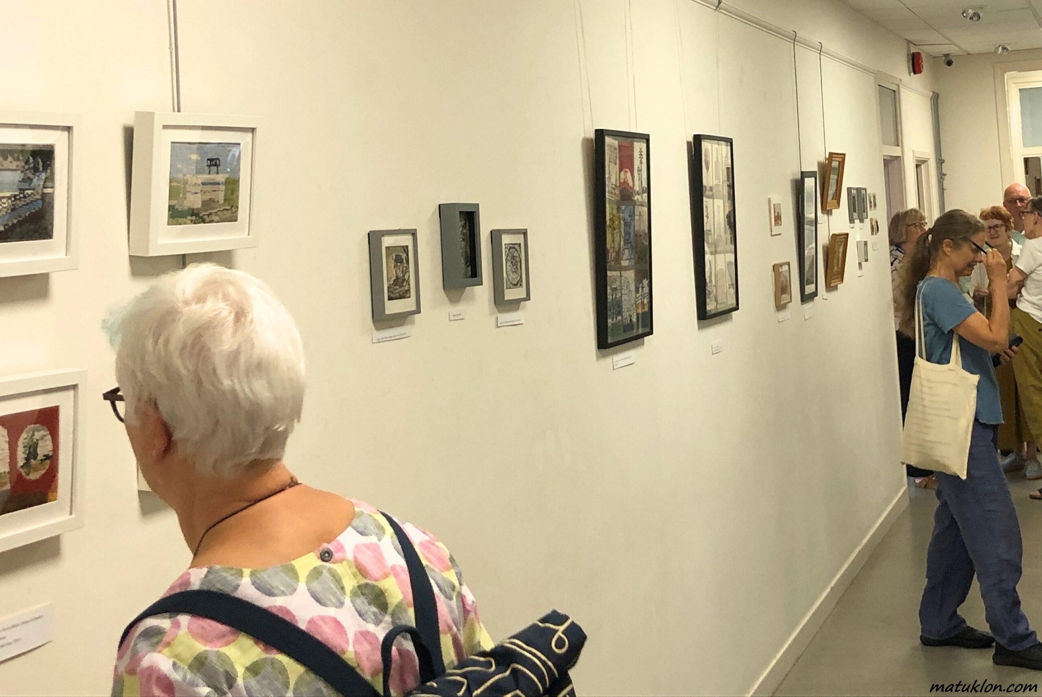 Woman looking at artworks on a white wall in a gallery; a group of people standing in the background