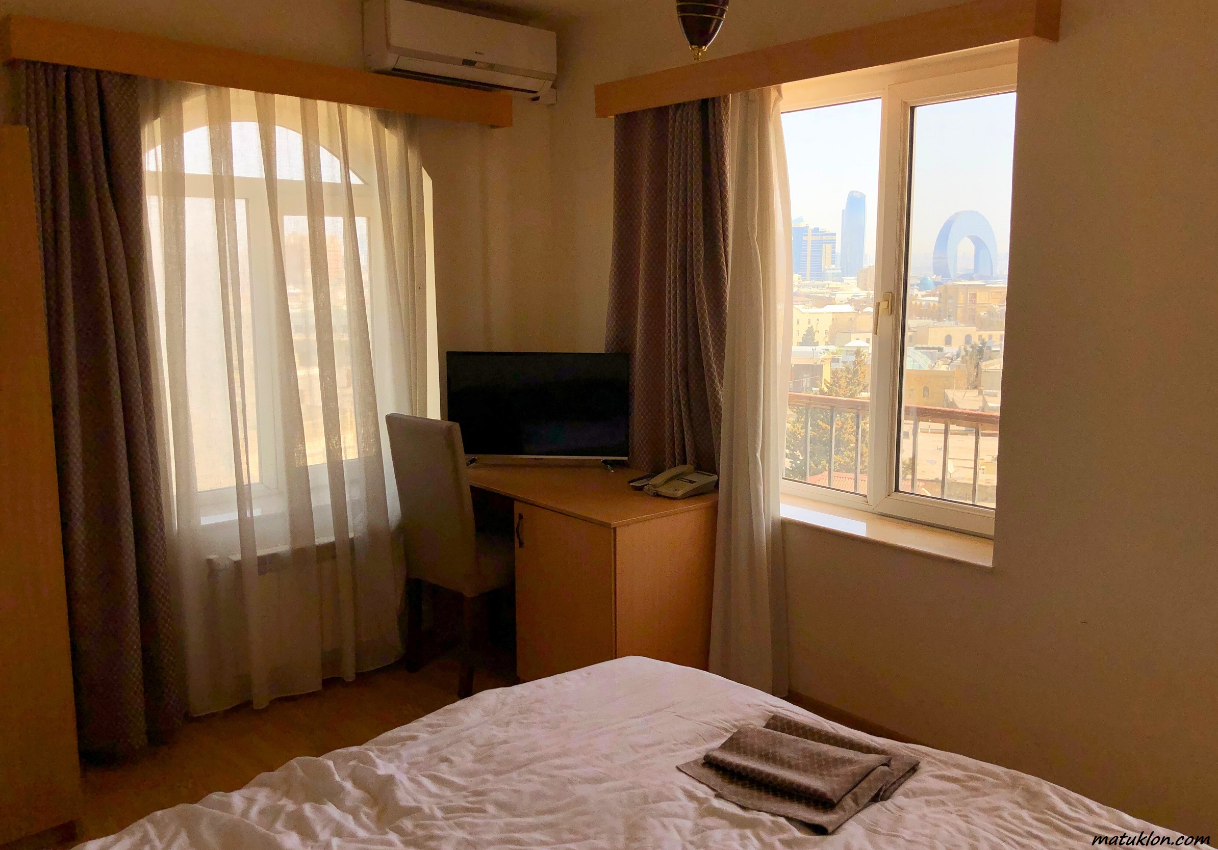 Hotel room; bed in the foreground, desk with TV set in the background; big windows in both walls