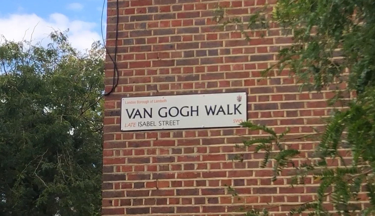 Street name plate saying "Van Gogh Walk, late Isabel Street" on a brick wall surrounded by greenery, with blue sky and white clouds in the background