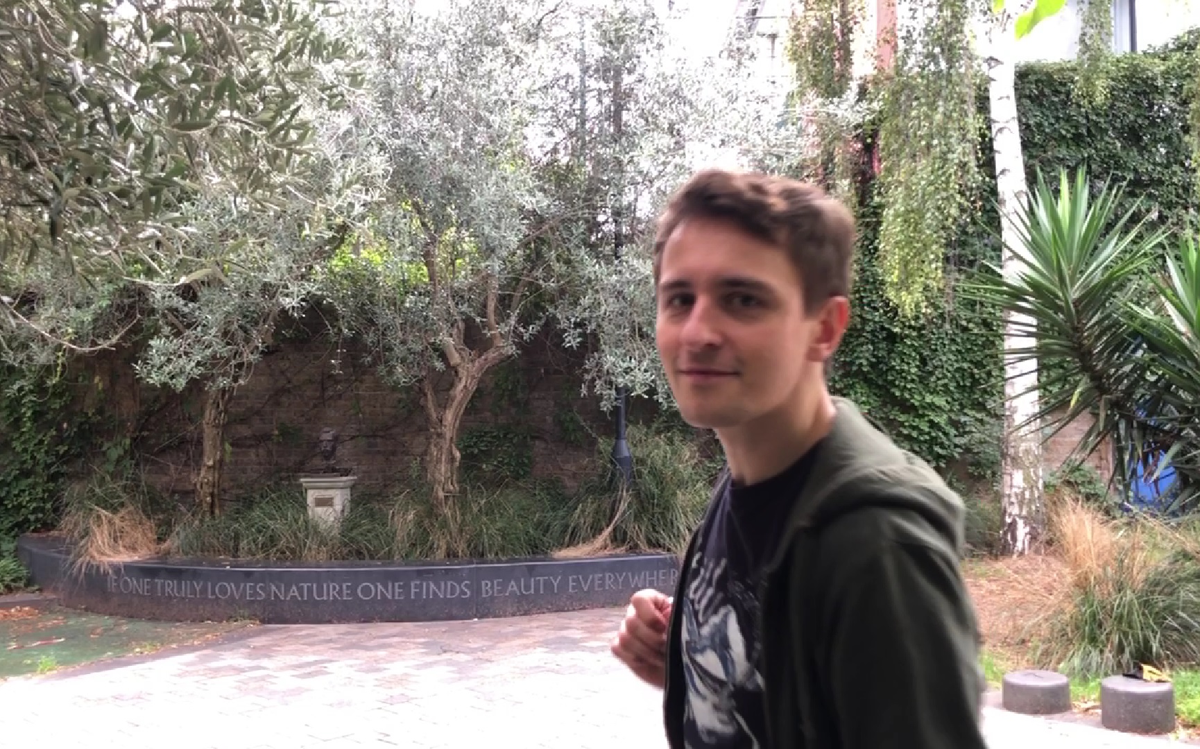 Man in green jacket standing sideways to the camera; greenery and a Vincent Van Gogh bust in the background